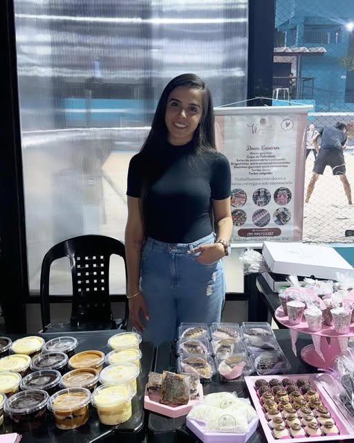 A confeiteira da Vi Doces sorrindo atrás da mesa de doces, com cupcakes, copos de sobremesa e brigadeiros
